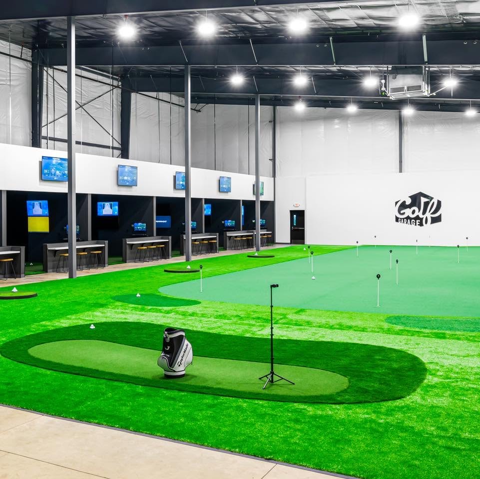 Interior of the Golf Garage indoor golf simulator facility, with putting green turf, hitting bays, and large screens showing virtual courses.
