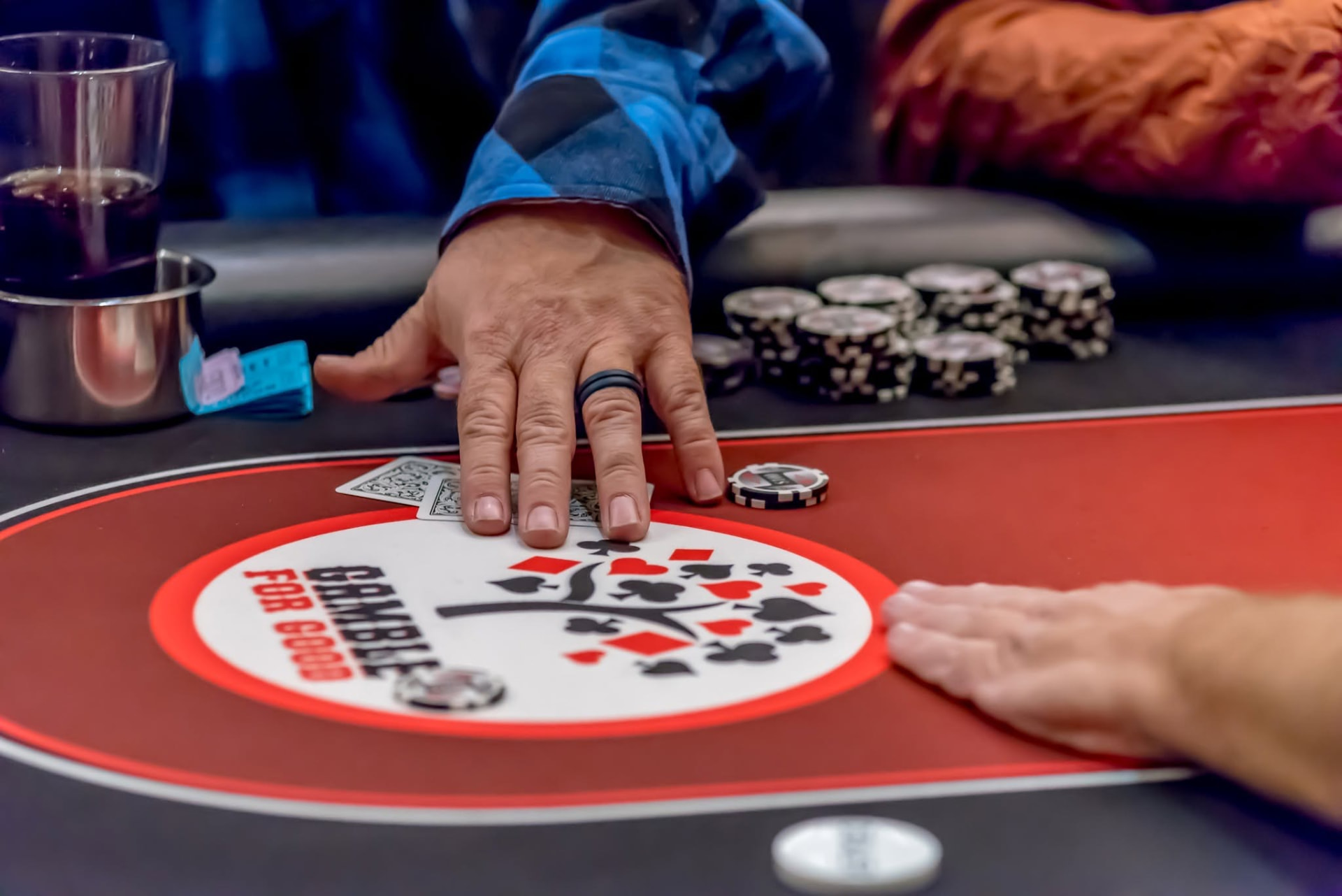 A hand resting on two face-down playing cards at the Gamerosity Belfor Poker Tournament, with poker chips and a Gamble For Good logo on the red felt table.